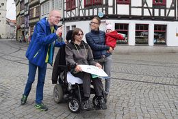 Zwei Männer und eine Frau mit Kind stehen am Domplatz. Ein Mann im Rollstuhl hat einen Stadtplan auf dem Schoß. Der andere Mann zeigt auf etwas.